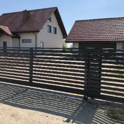 Nowoczesna, przesuwna brama z poziomych profili, w kolorze antracytowym, na tle domu z brązową dachówką. Cień bramy na piaszczystej drodze.