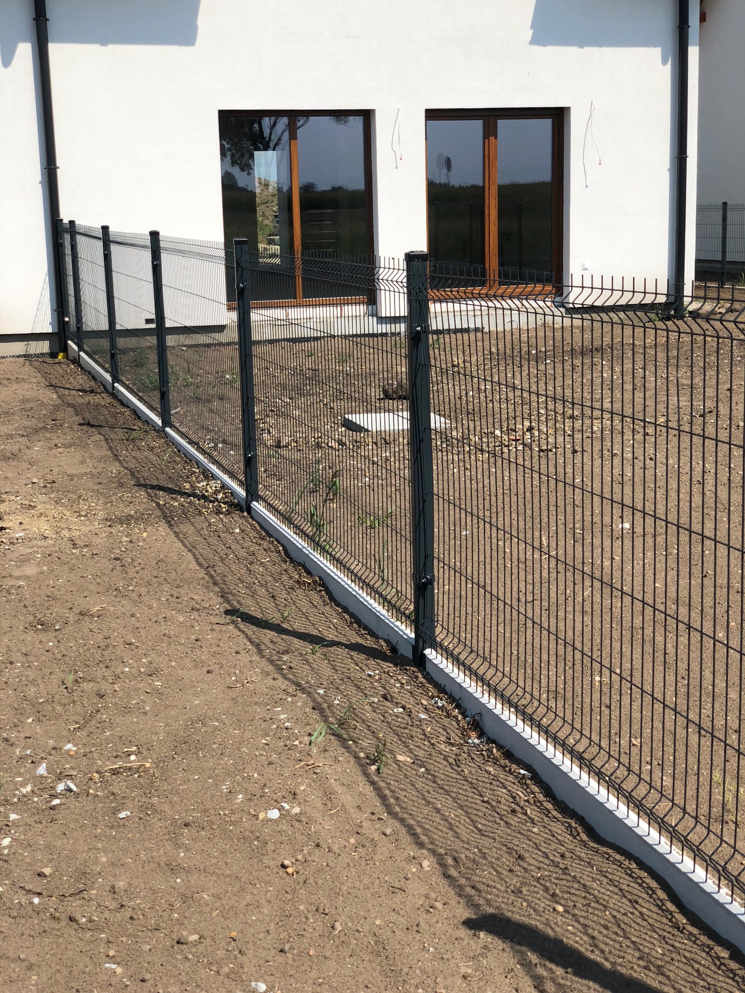 Nowoczesne ogrodzenie panelowe w kolorze grafitowym, zamontowane na betonowej podmurówce przy białym budynku z oknami. Widok z zewnątrz posesji, grunt w stanie surowym.