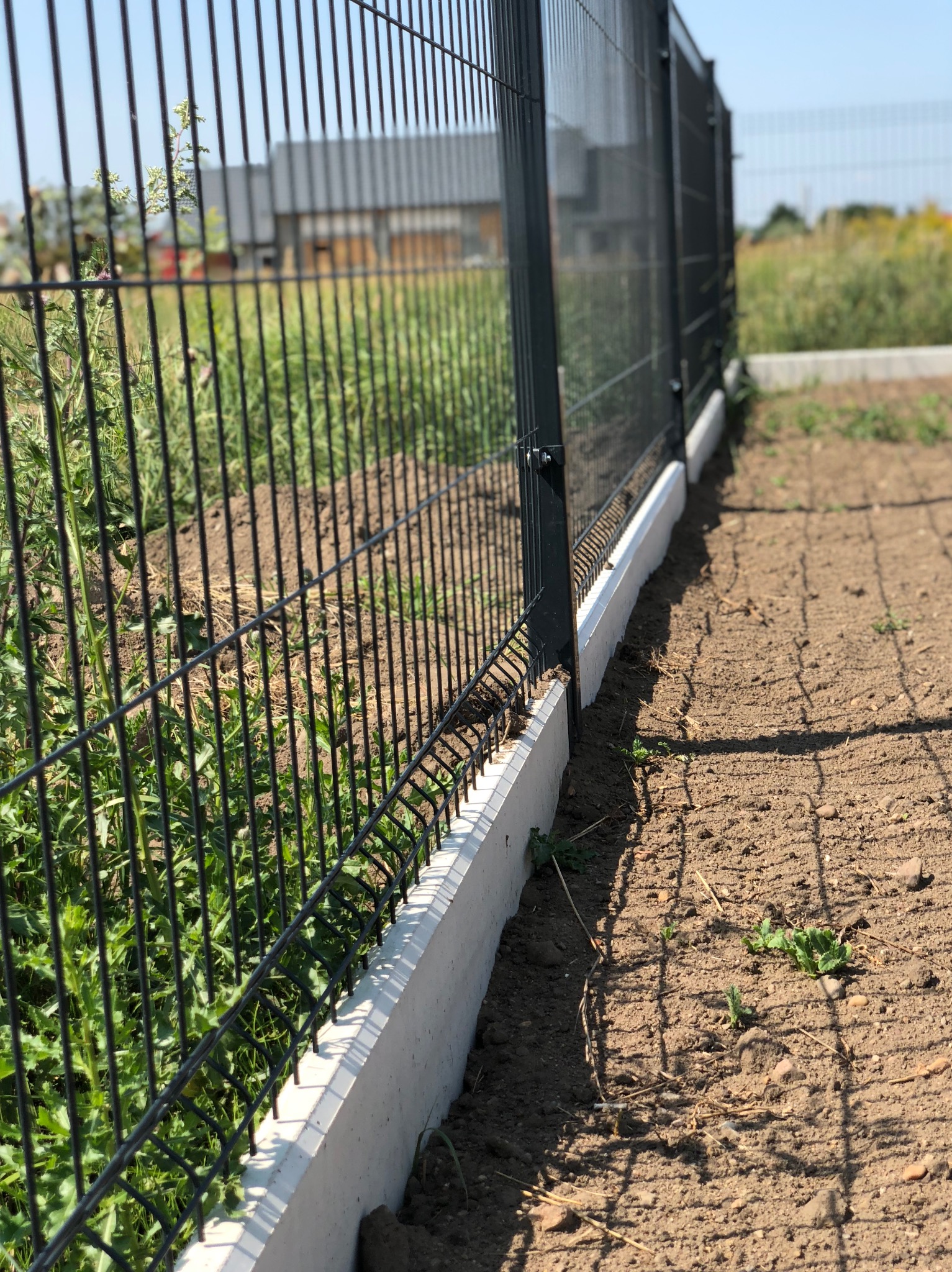 Nowoczesne, grafitowe ogrodzenie panelowe z podmurówką betonową, widoczne z bliska na tle zieleni i budynku w oddali. Kąt ujęcia podkreśla linię ogrodzenia.