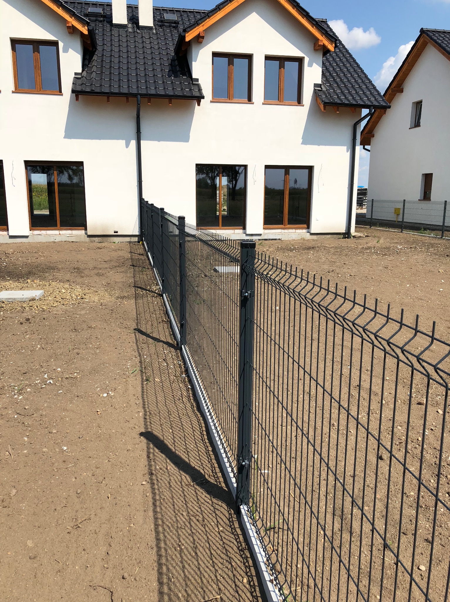 Nowoczesne, panelowe ogrodzenie w kolorze grafitowym, oddzielające posesję od sąsiedniego domu. Widoczne słupki i podmurówka. Tło stanowi biały dom z ciemną dachówką.