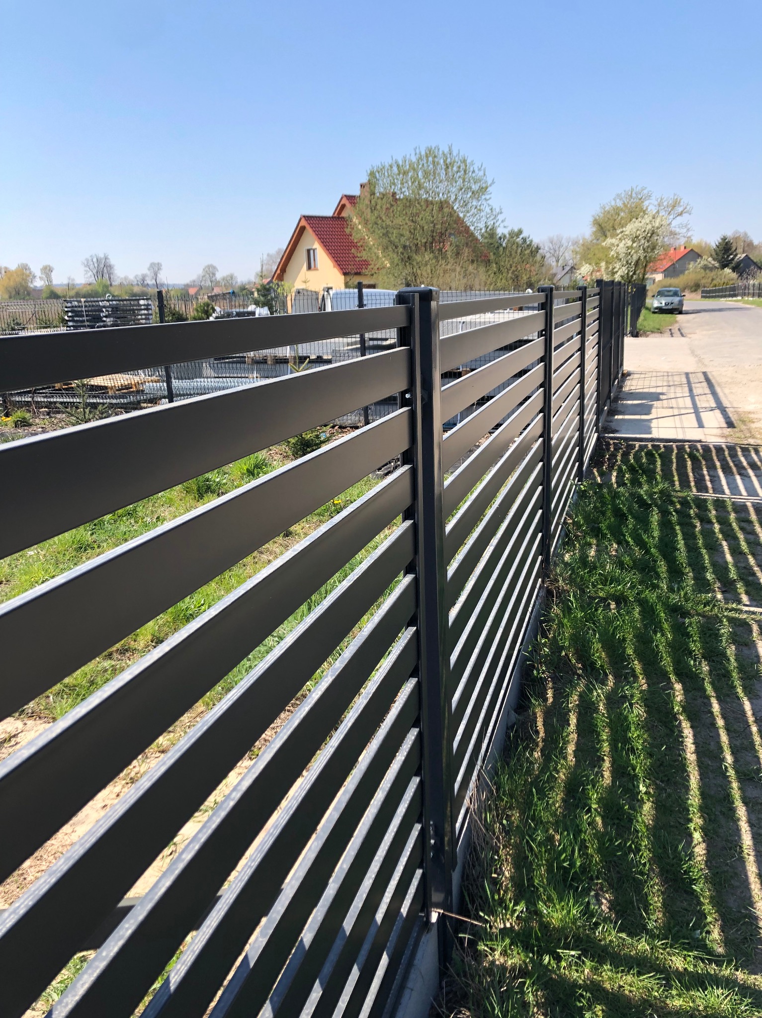 Nowoczesne, poziome przęsła ogrodzenia panelowego w kolorze grafitowym. Widok na dom z czerwoną dachówką i zielony trawnik. Cień ogrodzenia na trawie.