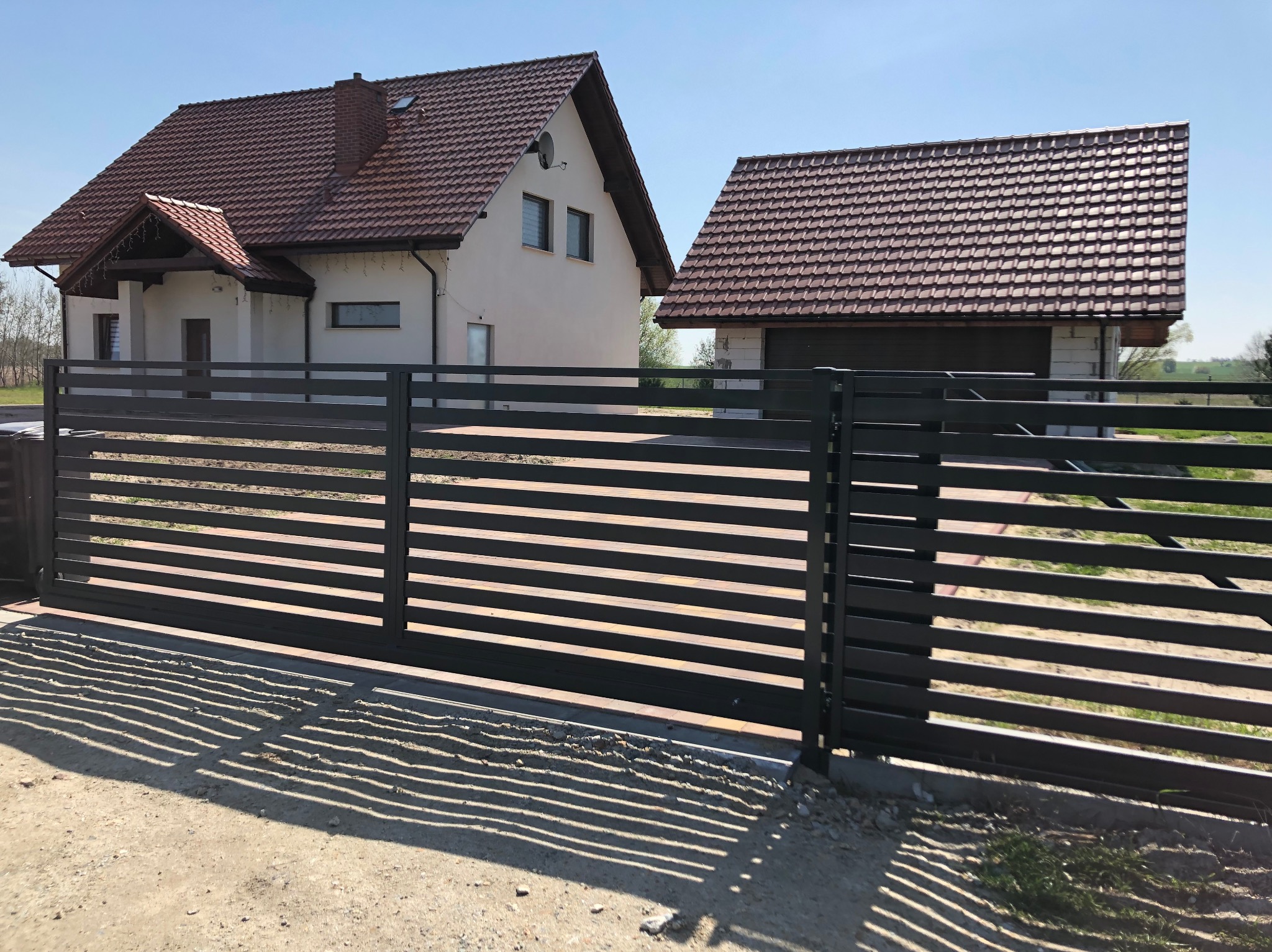 Nowoczesna, przesuwna brama z poziomych profili, w kolorze antracytowym, na tle domu z brązową dachówką. Cień bramy na piaszczystej drodze.