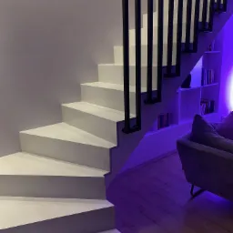 Nowoczesne, białe schody z czarną, minimalistyczną balustradą, oświetlone fioletowym światłem, z widocznym fragmentem regału z książkami i sofy w tle.
