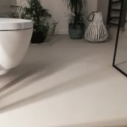 Beżowa, gładka posadzka w łazience z wiszącą toaletą, roślinami doniczkowymi i dekoracyjną latarnią, widoczna kabina prysznicowa z niebieskimi płytkami.