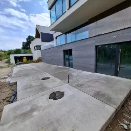 Nowoczesny dom z szarą elewacją i betonowym chodnikiem z włazami kanalizacyjnymi. Widoczne nieukończone prace ziemne i niebieskie rury. Pochmurne niebo.