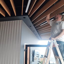 MG Project Grzegorz Wadas - Montaż drewnianej podbitki sufitowej przez pracownika na drabinie. Widoczne oświetlenie LED i wykończona ściana w pionowe pasy. Praca na wysokości w nowoczesnym wnętrzu.
