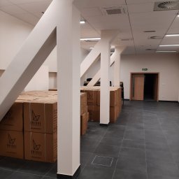 MG Project Grzegorz Wadas - Białe, pomalowane filary w biurze z szarą podłogą, z kartonami z logo 'Unique' w tle. Widoczny sufit z oświetleniem i systemem wentylacji. Otwarta przestrzeń biurowa.