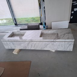 MG Project Grzegorz Wadas - Marmurowa szafka pod umywalkę z dwoma otworami, ustawiona na drewnianych klockach w surowym wnętrzu łazienki w trakcie wykończenia. Widoczne okno i fragmenty ścian.