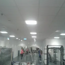 Jasne wnętrze siłowni w trakcie odświeżania: drabina, osoby pracujące, sprzęt do ćwiczeń. Sufit kasetonowy z oświetleniem. Widok na ścianę z lustrami.