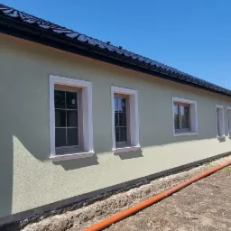 Jasnozielona elewacja domu jednorodzinnego z białymi obramowaniami okien, czarną dachówką i rurą spustową, widoczna instalacja w wykopie wzdłuż fundamentów budynku.