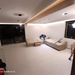 Wnętrze po remoncie z gładkimi, pomalowanymi na biało ścianami i sufitem, z meblami zabezpieczonymi folią, widocznym oświetleniem LED w suficie i lampą wiszącą.