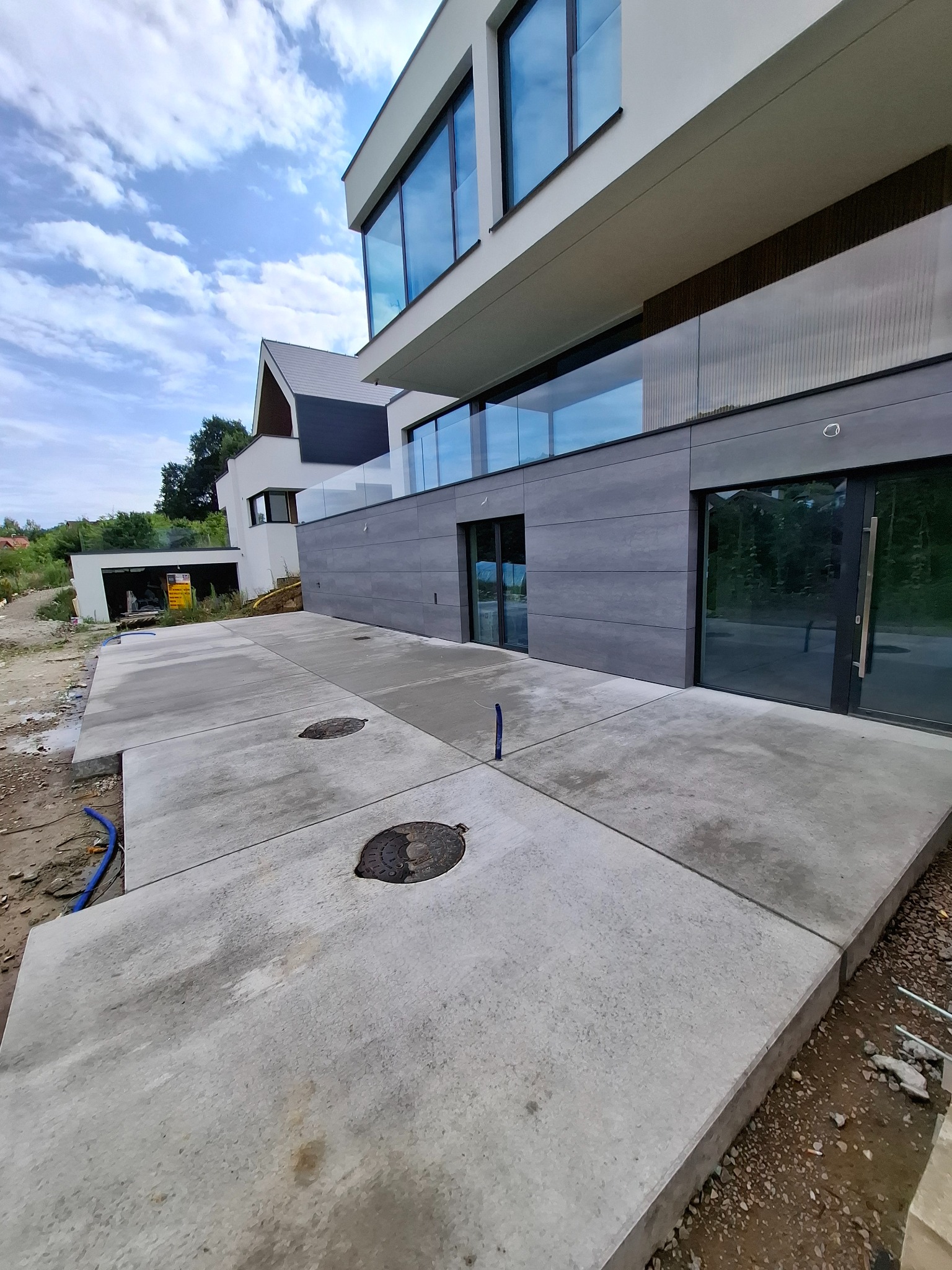 Nowoczesny dom z szarą elewacją i betonowym chodnikiem z włazami kanalizacyjnymi. Widoczne nieukończone prace ziemne i niebieskie rury. Pochmurne niebo.