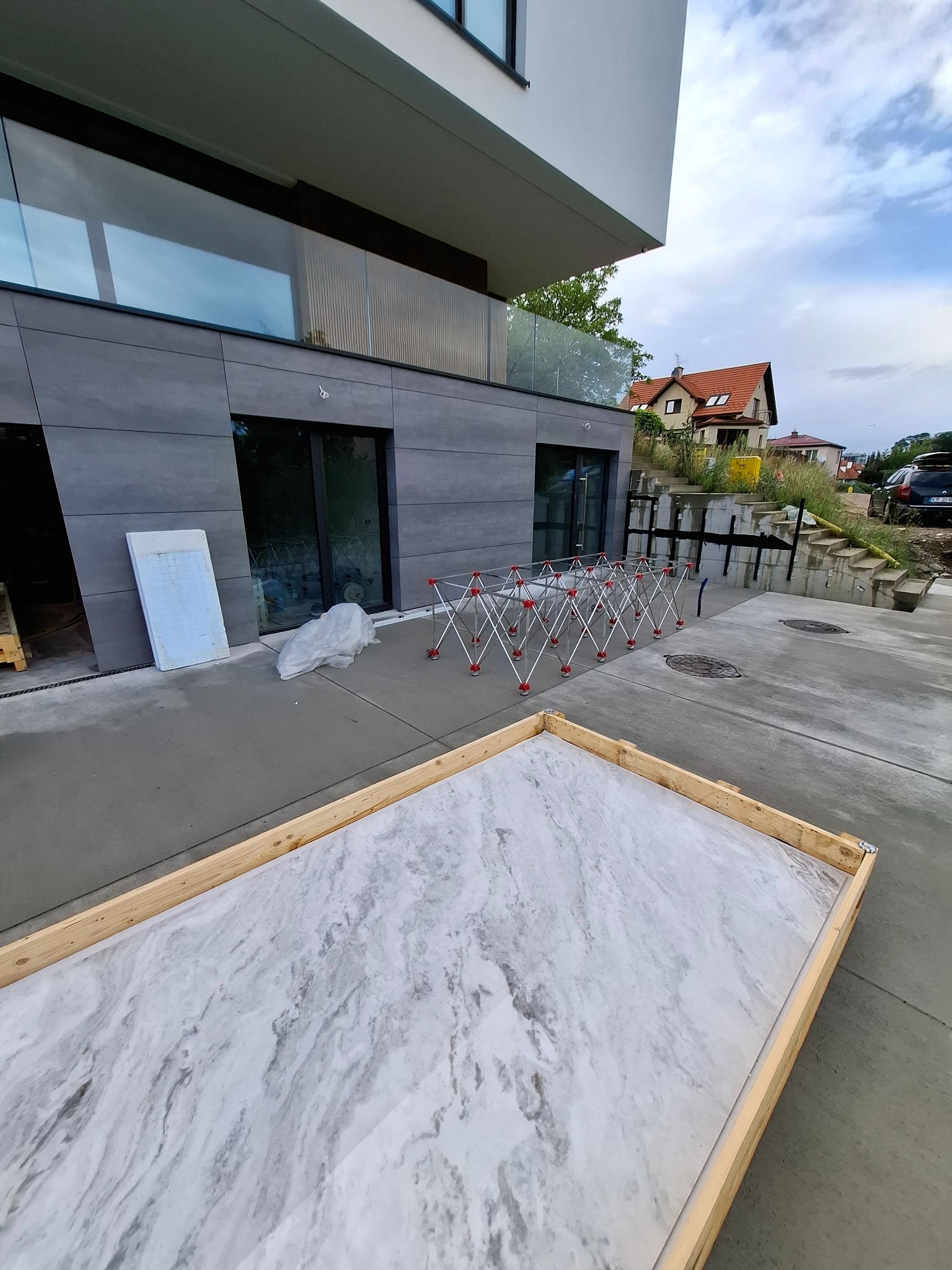 Płyta z marmurowym wzorem w drewnianej ramie na betonowym podłożu przed budynkiem z szarymi panelami. W tle dom jednorodzinny i niebo z chmurami.