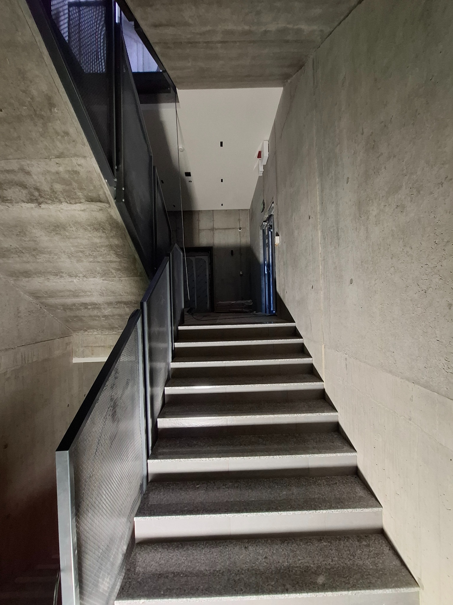 Betonowe schody z metalową balustradą w surowym, nowoczesnym wnętrzu. Widoczna faktura betonu na ścianach i suficie. Minimalistyczne oświetlenie.