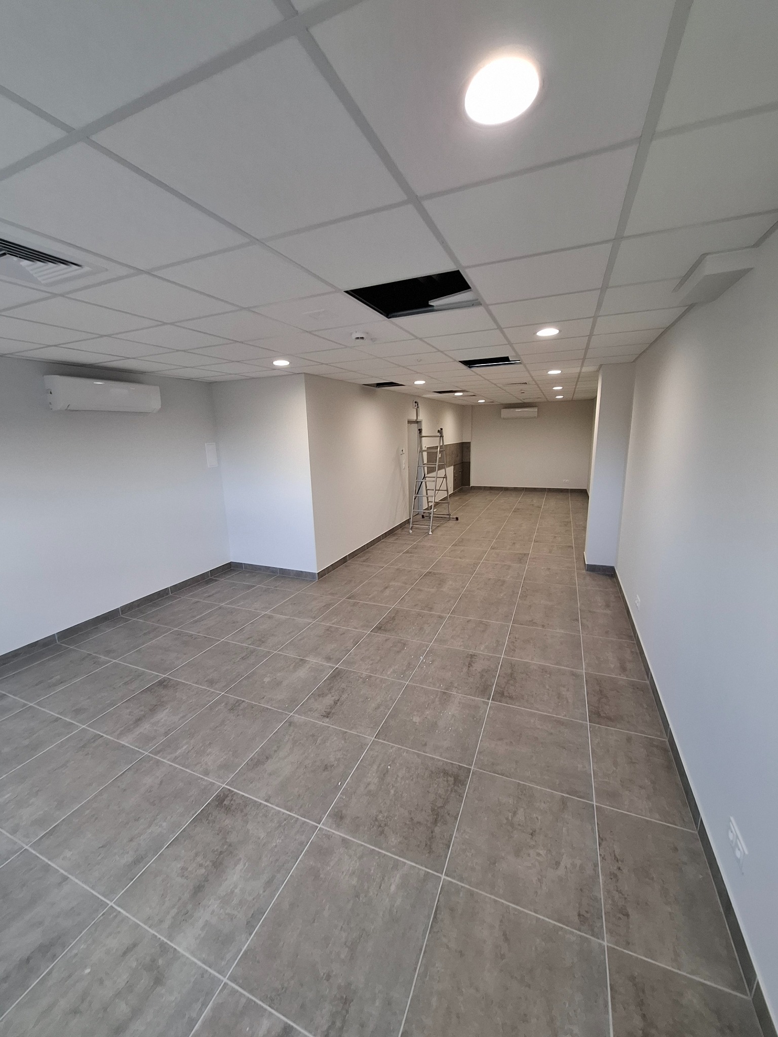 Wykończone, puste pomieszczenie biurowe z szarą podłogą z płytek, białymi ścianami i sufitem kasetonowym z widoczną instalacją. W tle drabina.