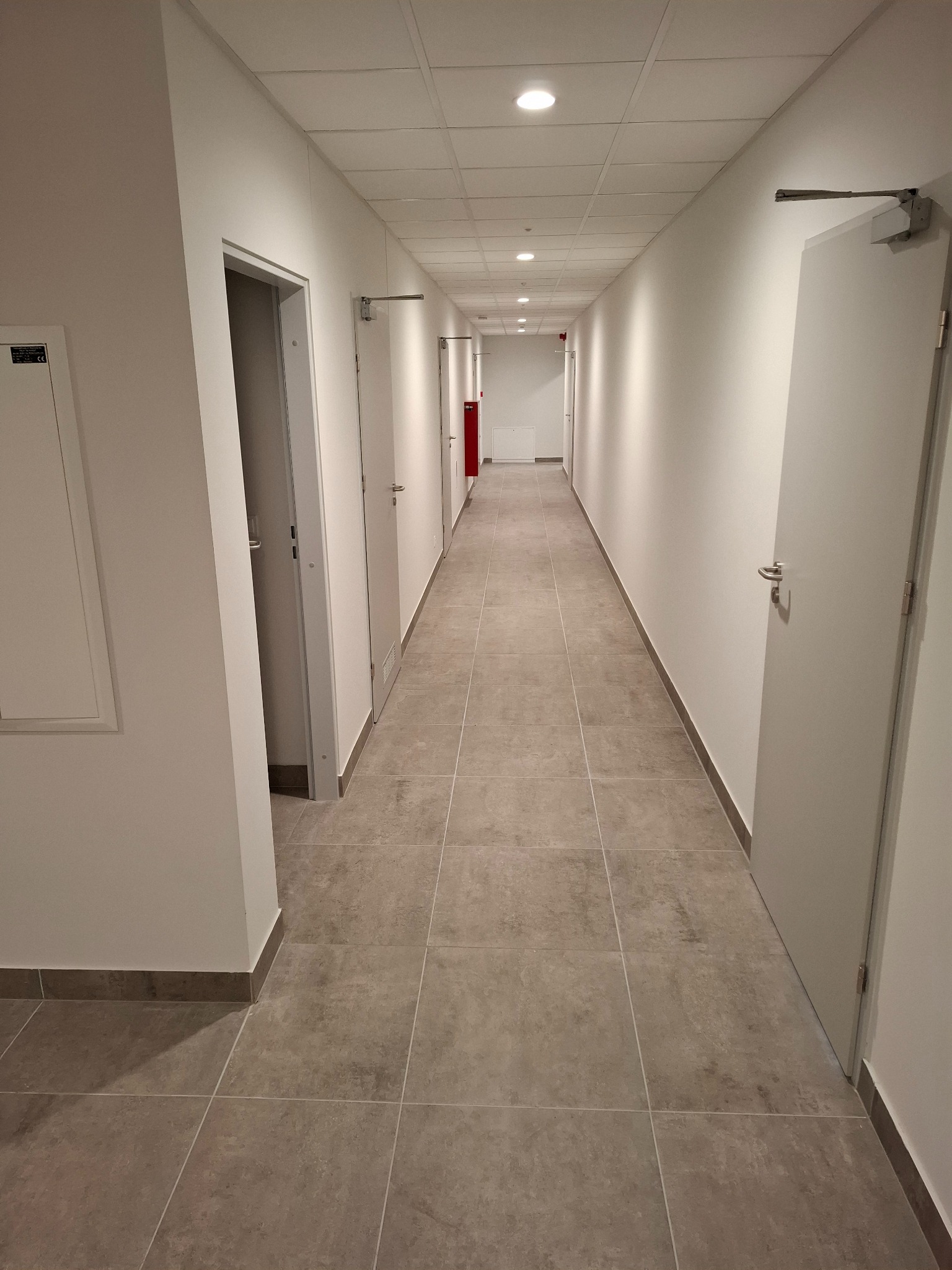 Długi, jasny korytarz z szarymi płytkami podłogowymi i białymi ścianami z drzwiami. Widoczne oświetlenie sufitowe i detale wykończenia.