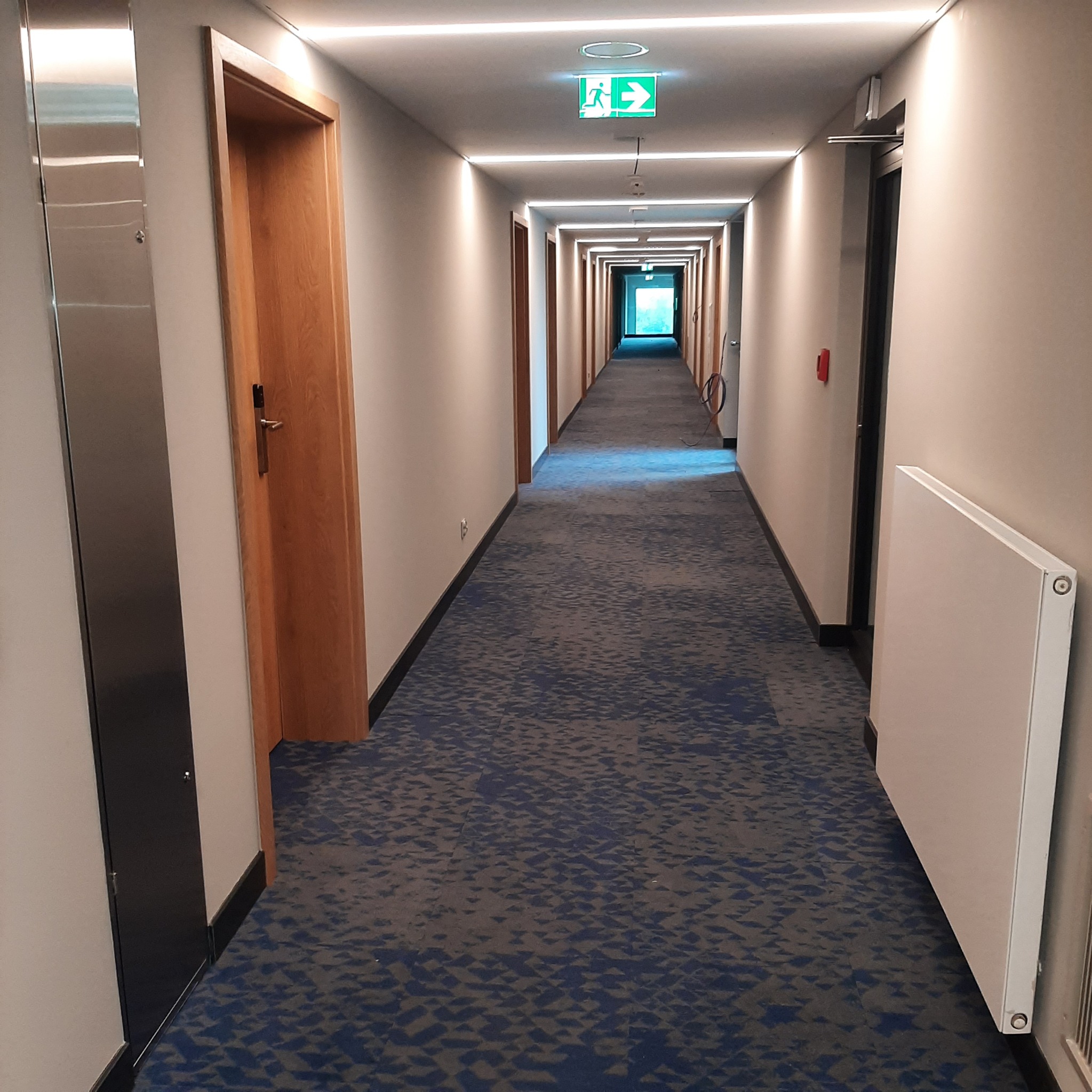 Długi korytarz z niebieską wykładziną i oświetleniem LED. Widoczne drzwi, grzejnik i znak ewakuacyjny. Perspektywa zbieżna w kierunku światła na końcu korytarza.