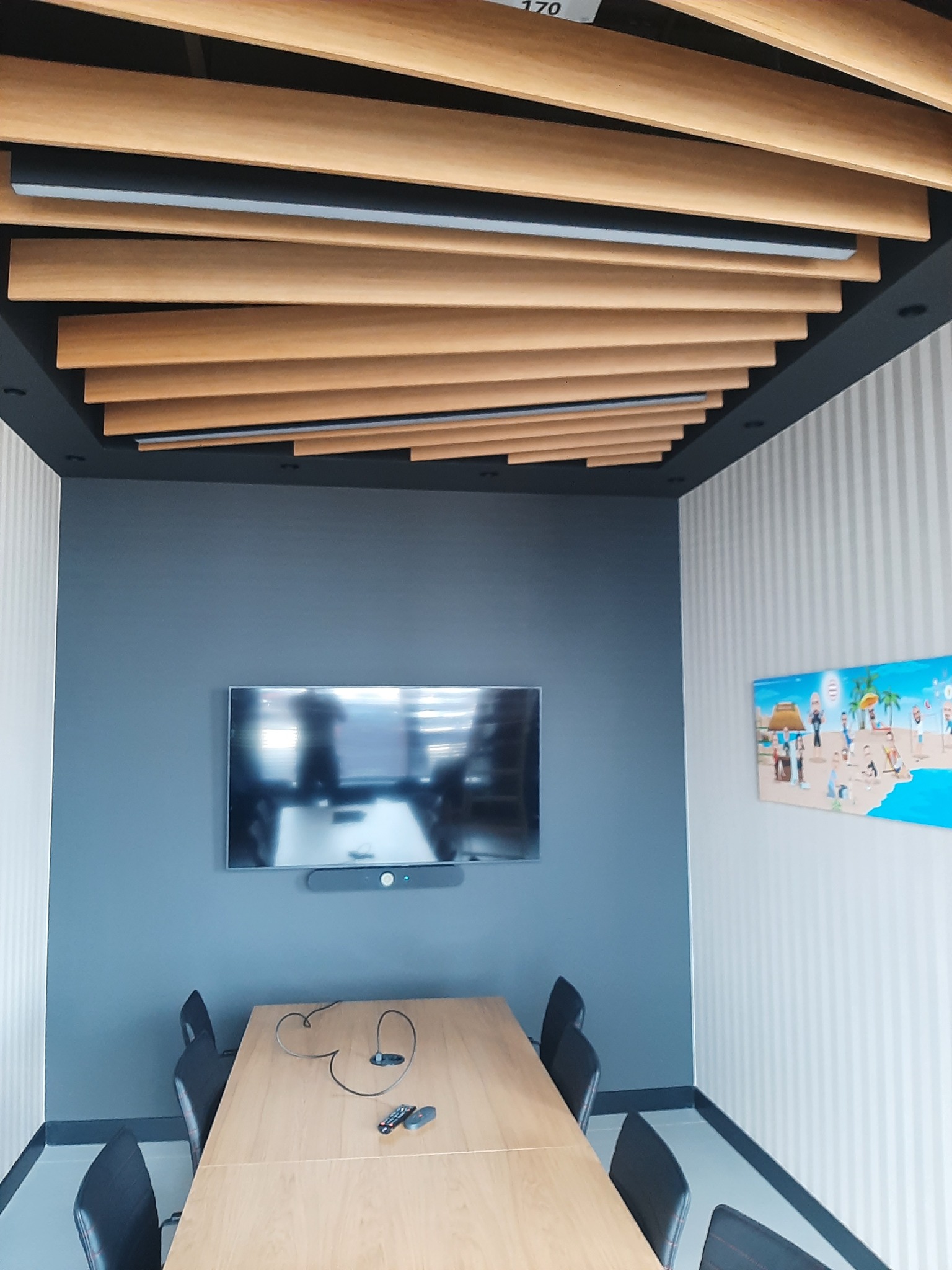 Nowoczesna sala konferencyjna z drewnianym sufitem lamelowym, szarą ścianą z telewizorem i stołem z krzesłami. Ściana w paski i obraz dodają charakteru.