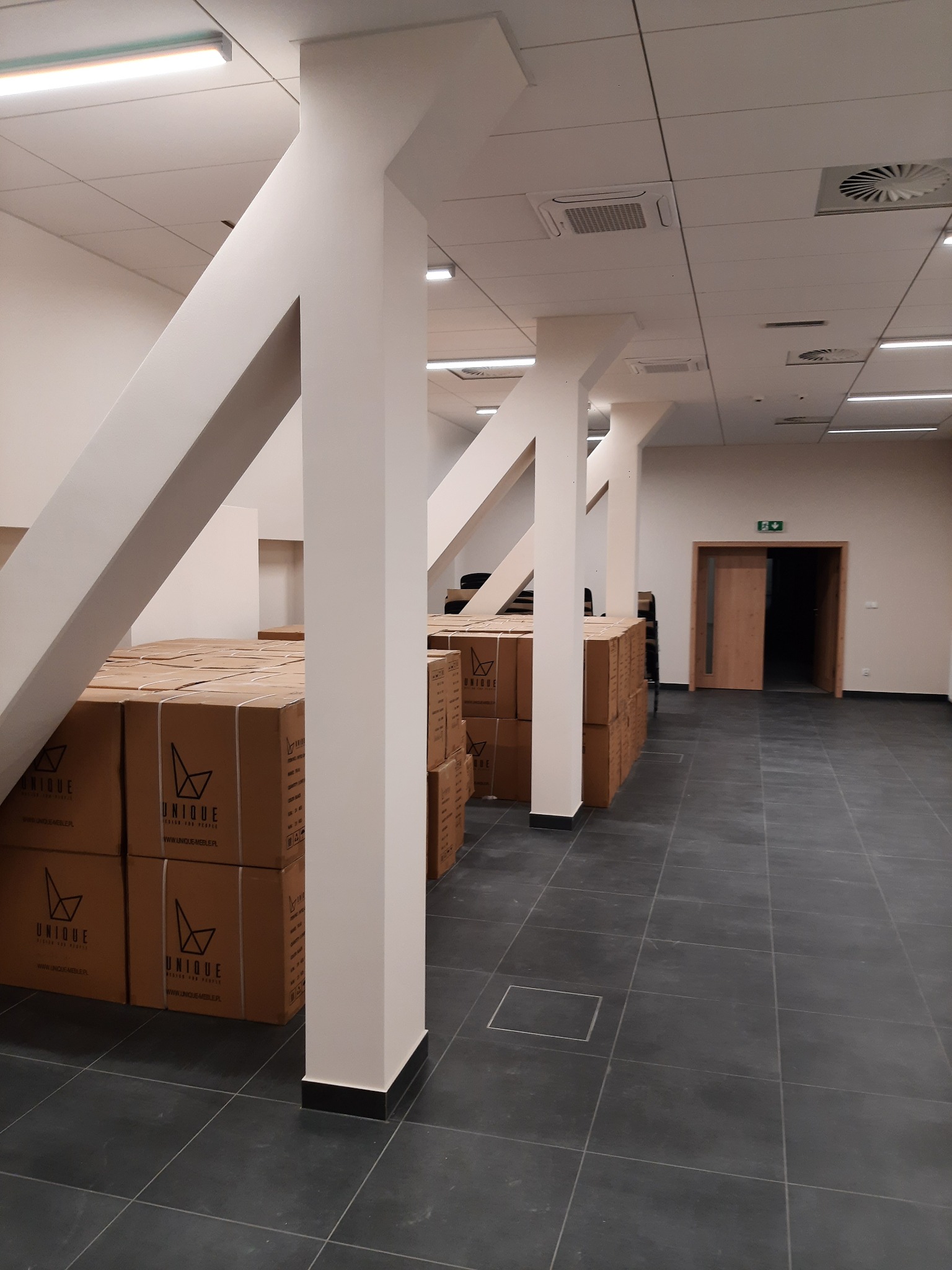 Białe, pomalowane filary w biurze z szarą podłogą, z kartonami z logo 'Unique' w tle. Widoczny sufit z oświetleniem i systemem wentylacji. Otwarta przestrzeń biurowa.