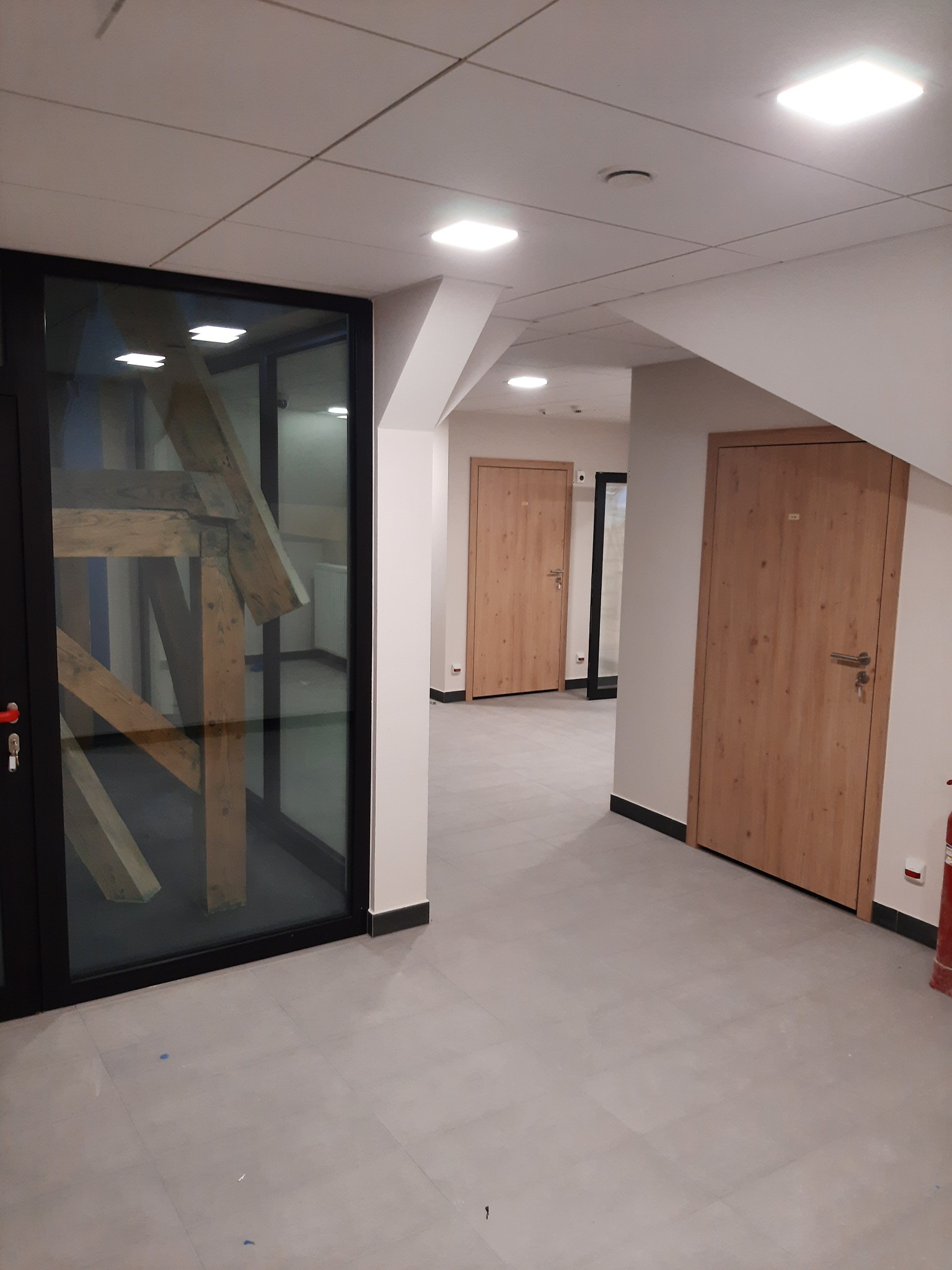 Korytarz biurowy z drewnianymi drzwiami i szklaną ścianą z widoczną konstrukcją. Jasne wnętrze, minimalistyczny design, płytki podłogowe w odcieniach szarości.