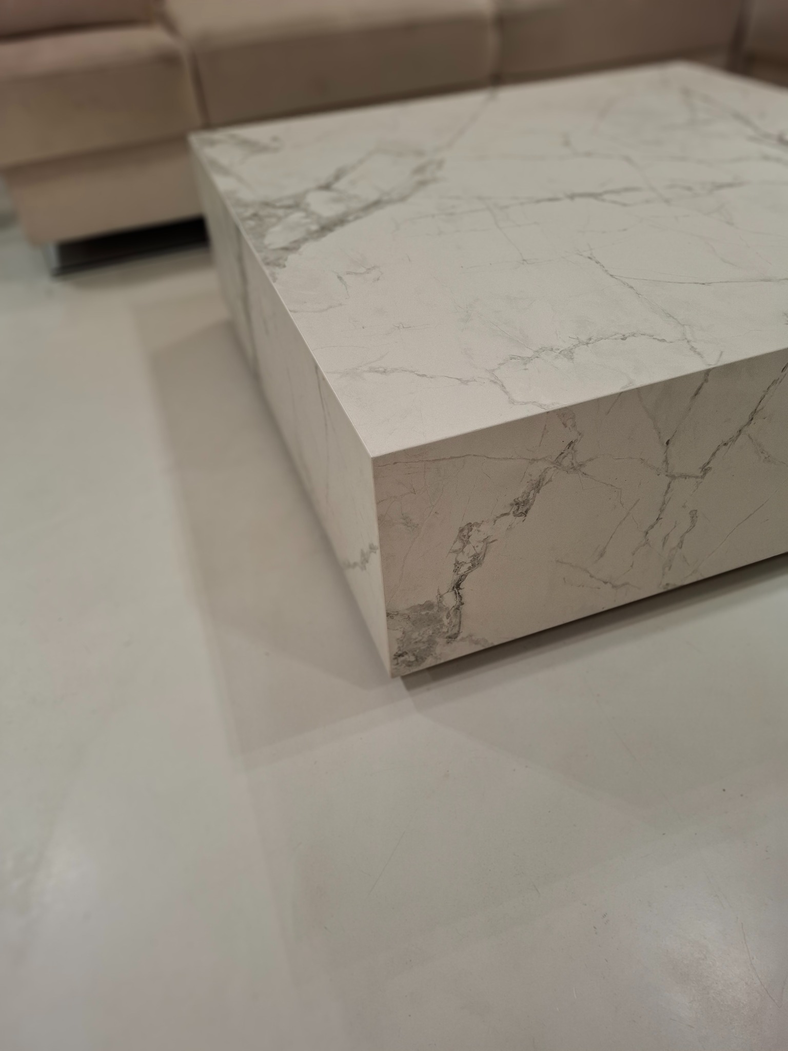 Kanciasty stolik kawowy z białego marmuru z szarymi żyłkami, ustawiony na jasnej podłodze, obok beżowej sofy. Minimalistyczny design.