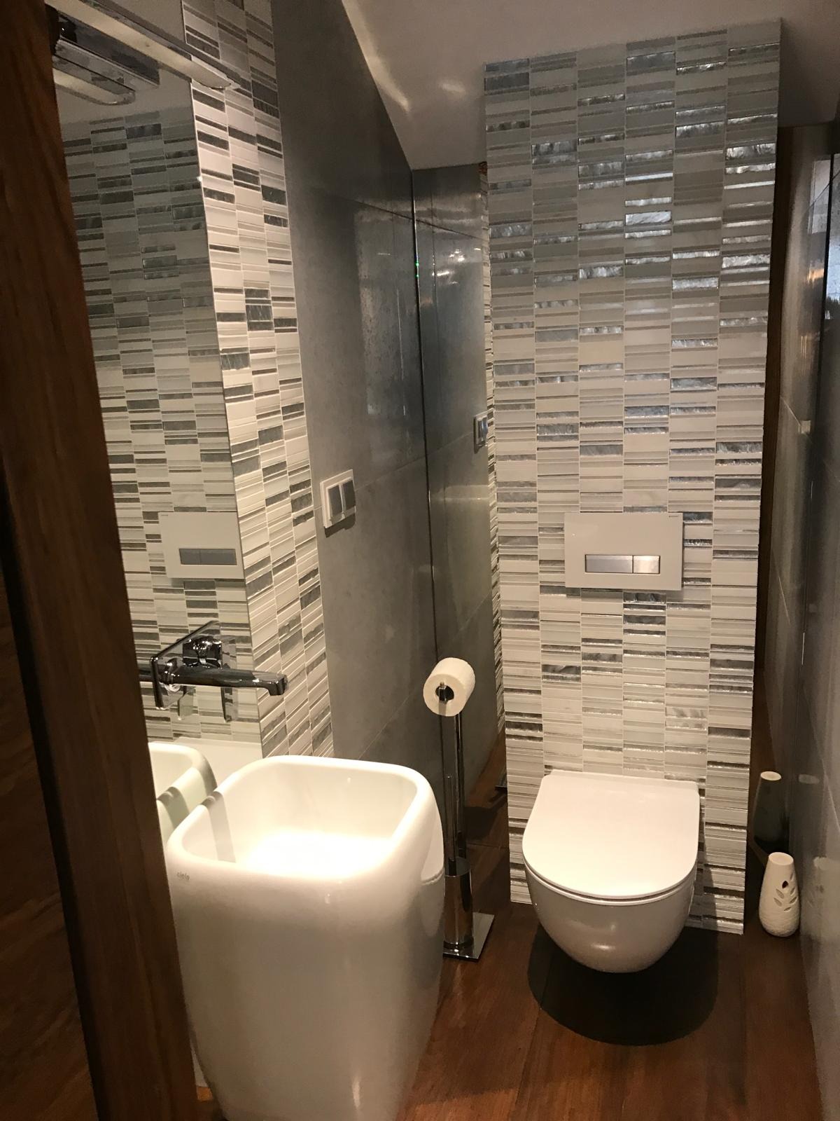 Nowoczesna łazienka z mozaiką w odcieniach szarości, białą umywalką na postumencie i podwieszaną toaletą. Drewniana podłoga dodaje ciepła minimalistycznemu wnętrzu.