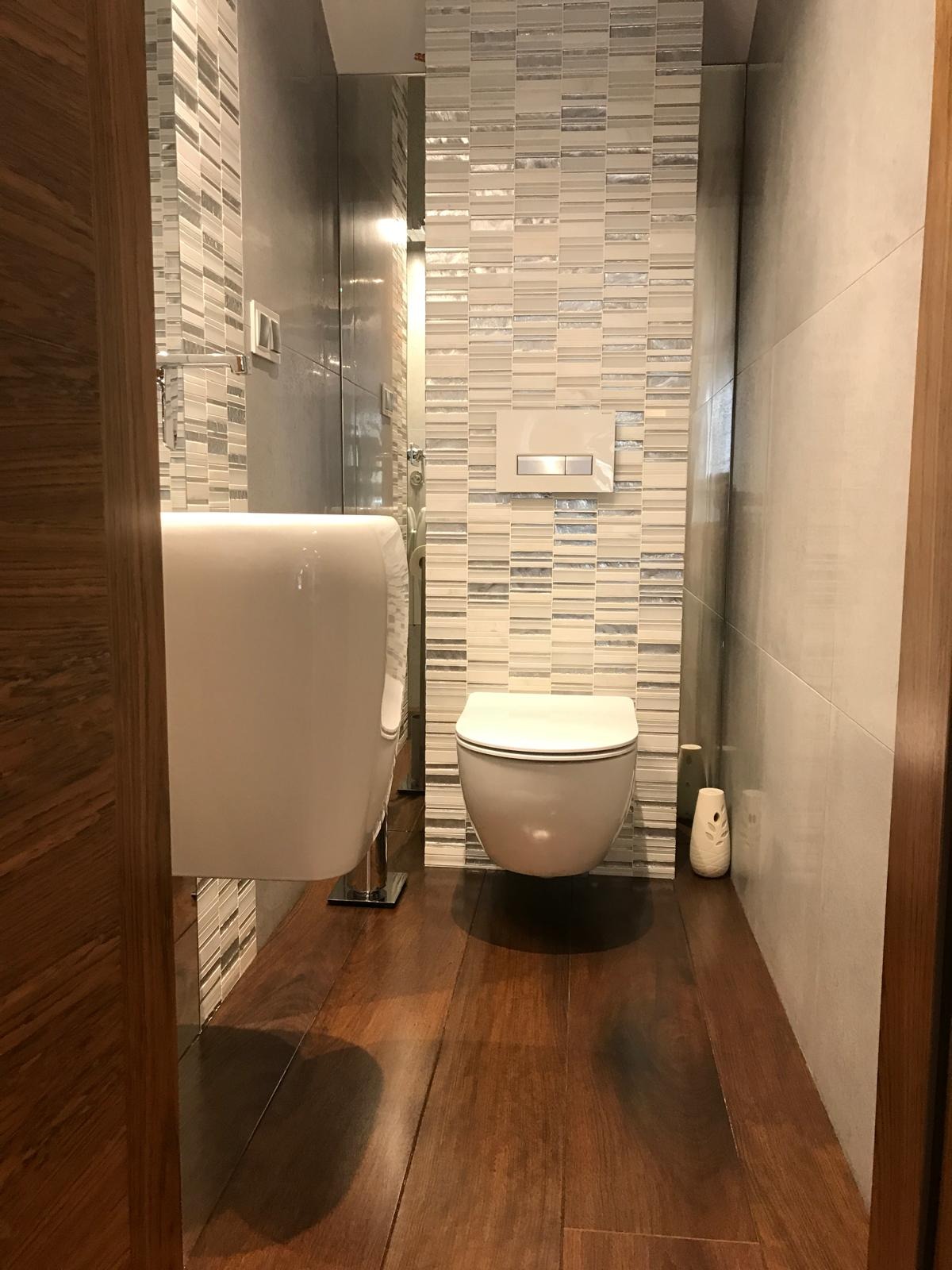 Nowoczesna aranżacja łazienki w Babicach: podwieszana toaleta, umywalka na tle mozaiki i drewniana podłoga. Minimalistyczny design, eleganckie wykończenie.