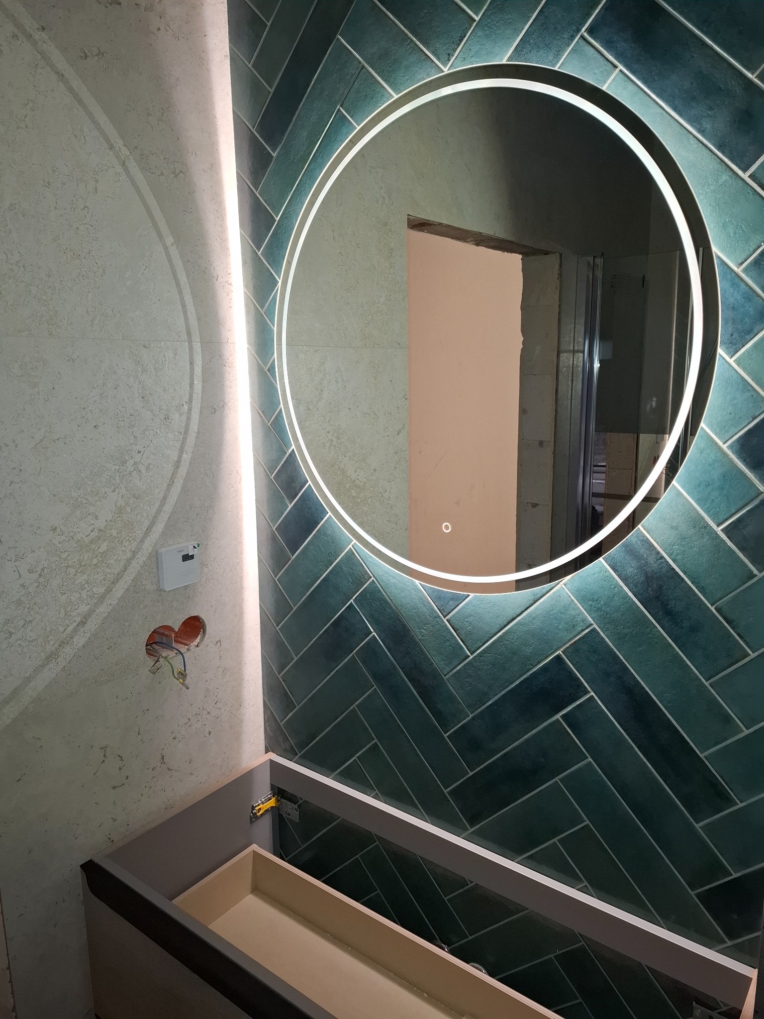 Niebiesko-zielona ściana z płytek ułożonych w jodełkę, okrągłe lustro z podświetleniem LED, niedokończona szafka pod umywalkę i wystające kable elektryczne.