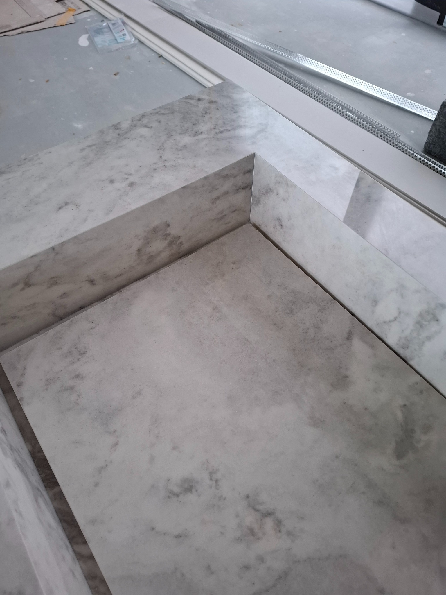 Wykończony blat łazienkowy z szarego marmuru, widoczny z góry, z metalowym profilem przy ścianie. Minimalistyczny design, surowe wnętrze w trakcie prac wykończeniowych.