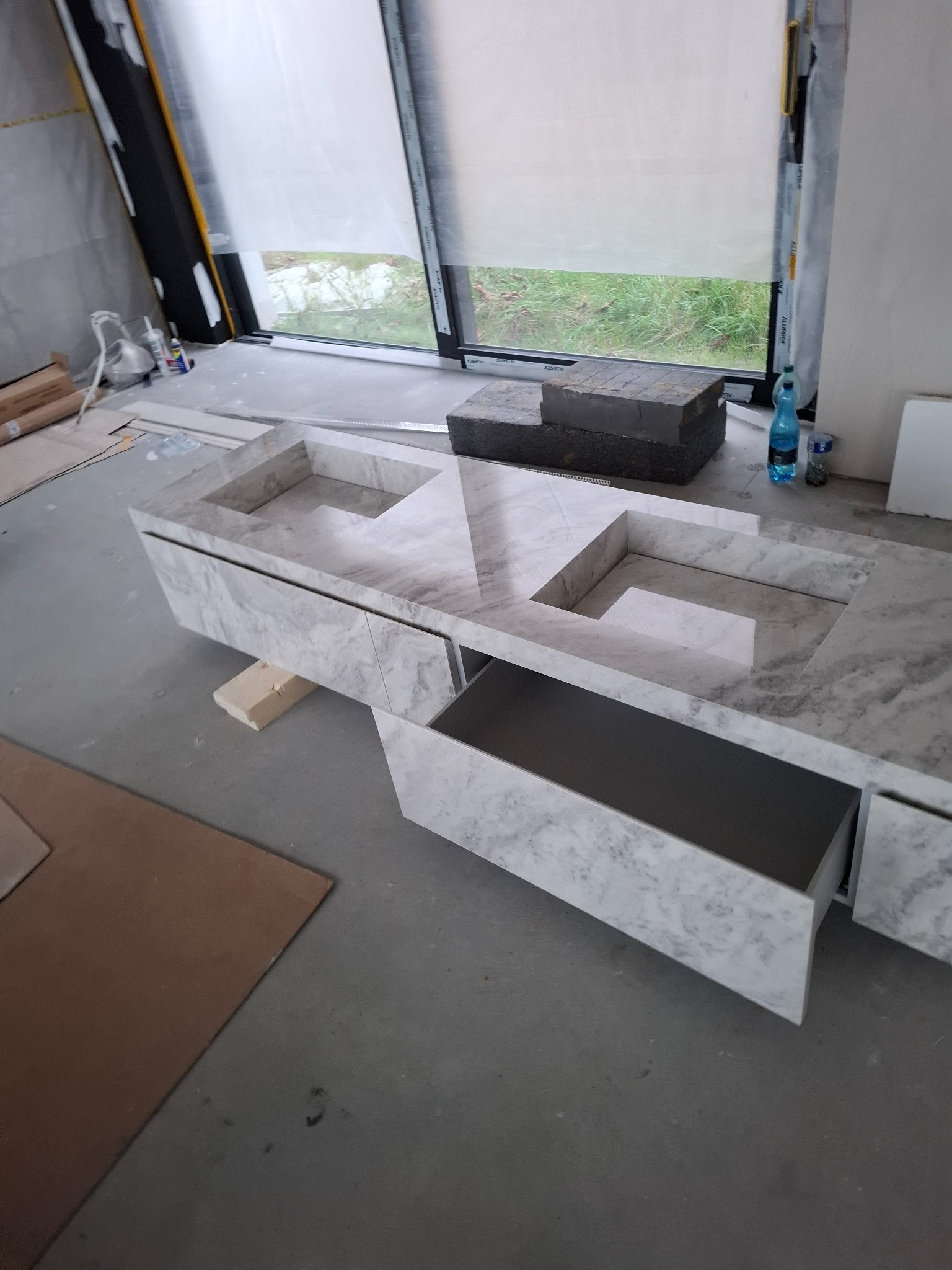 Marmurowa umywalka z szafką w łazience w trakcie wykańczania, z otwartą szufladą i widocznym oknem w tle. Umywalka z dwoma komorami.