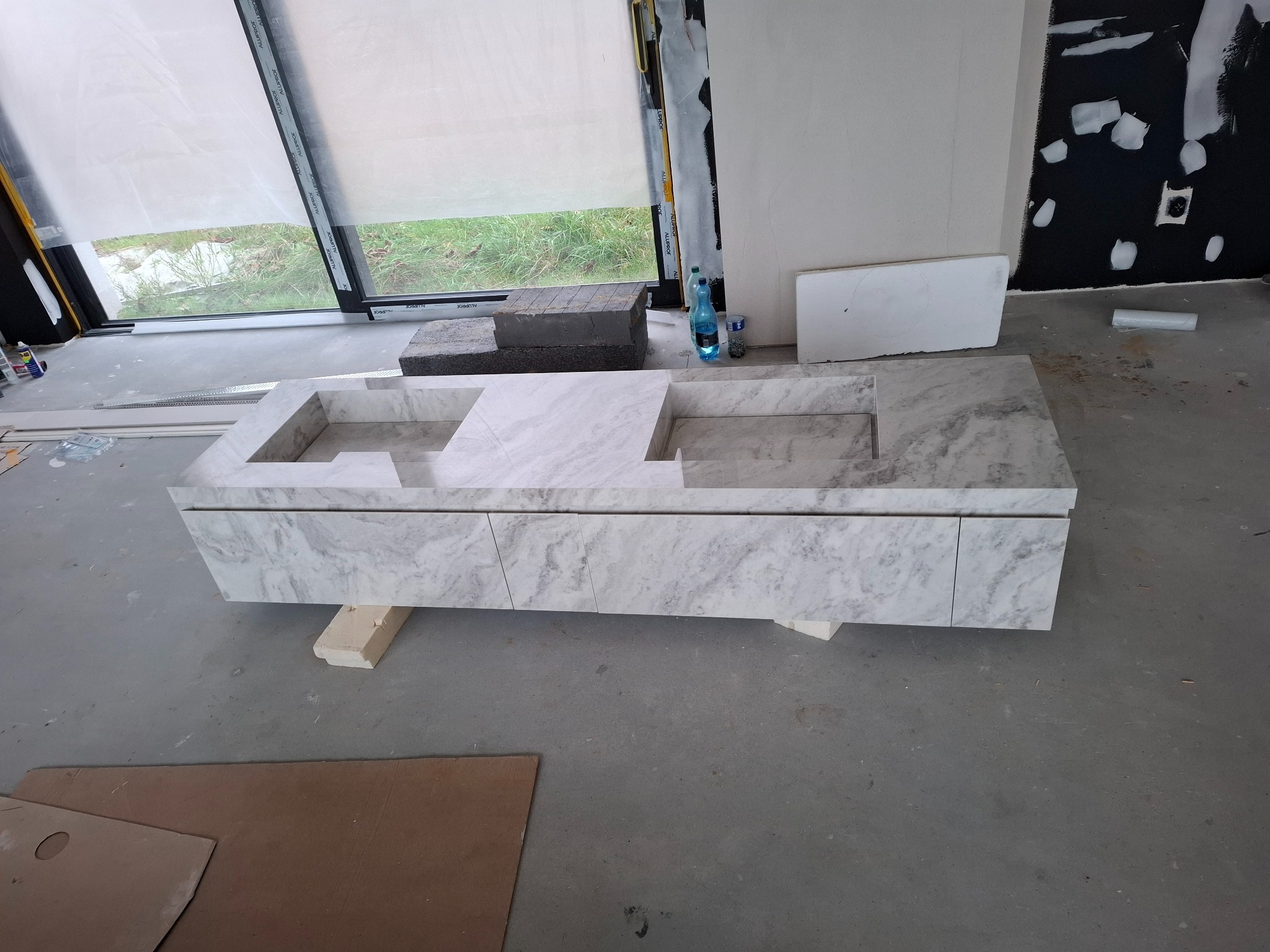 Marmurowa szafka pod umywalkę z dwoma otworami, ustawiona na drewnianych klockach w surowym wnętrzu łazienki w trakcie wykończenia. Widoczne okno i fragmenty ścian.