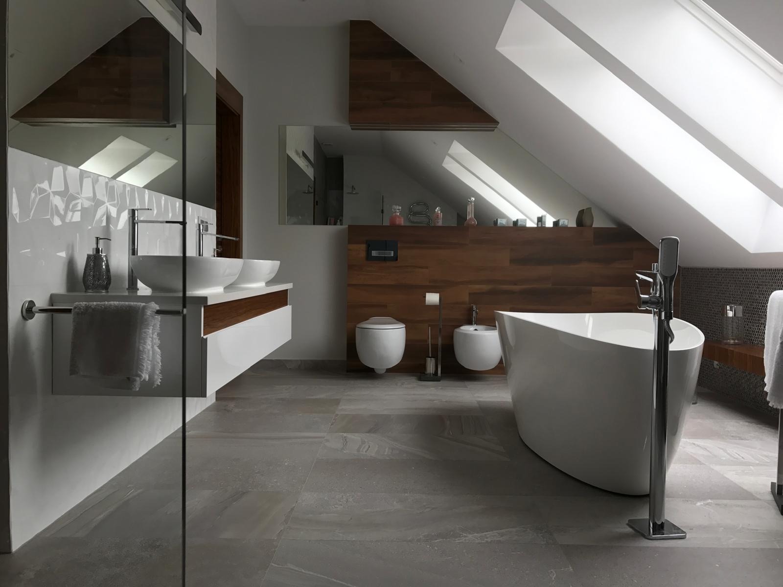 Nowoczesna łazienka na poddaszu z drewnianymi akcentami, wolnostojącą wanną i umywalkami nablatowymi. Uwagę przykuwają okna dachowe wpuszczające naturalne światło.