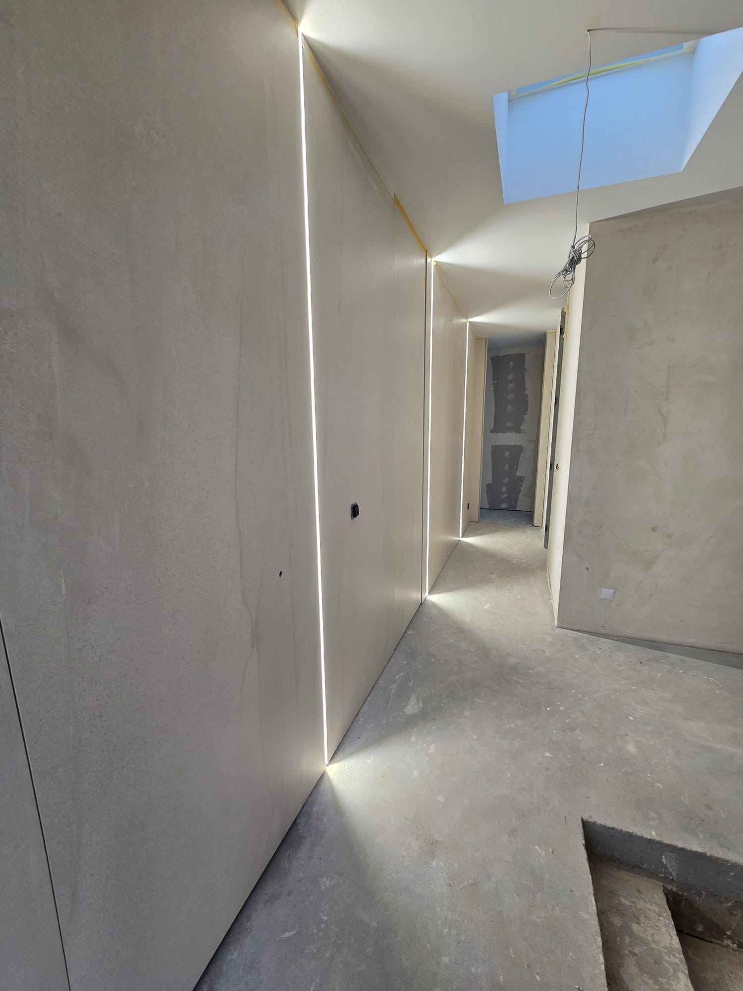 Wnętrze w trakcie wykańczania: szpachlowane ściany z wbudowanym oświetleniem LED, surowa podłoga, widok na kolejne pomieszczenia. Skąpe światło dzienne z okna dachowego.
