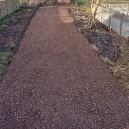 Ciemnobrązowa, żwirowa droga w trakcie budowy, z widocznym stosem ziemi w tle i fragmentami ogrodzenia po bokach.