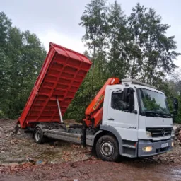 Biała ciężarówka z czerwoną, podniesioną skrzynią załadowaną gruzem ceglanym, stojąca na błotnistym gruncie w otoczeniu drzew.