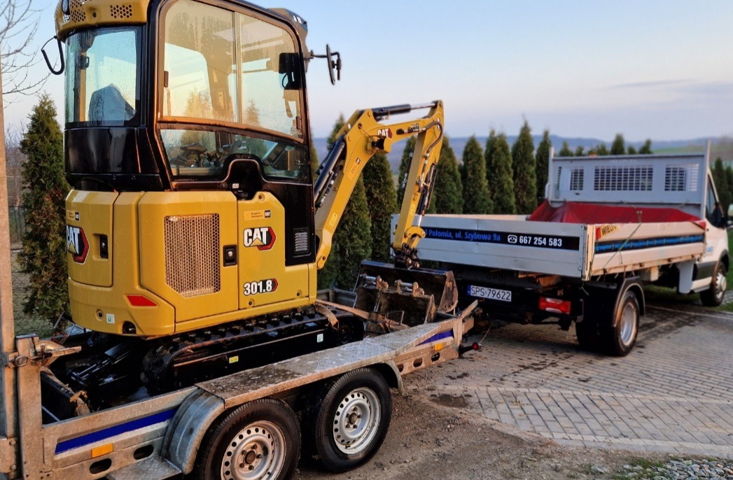 Żółta minikoparka CAT 301.8, załadowana na niskopodwoziową przyczepę, ciągniętą przez biały samochód dostawczy z wywrotką, na tle zielonych drzew i błękitnego nieba.