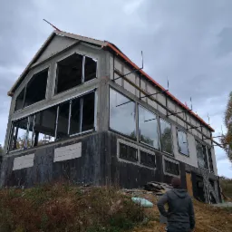 Surowy stan dwupiętrowego budynku z zamontowanymi oknami, widoczne rusztowania i przygotowania do dalszych prac wykończeniowych, osoba obserwująca postęp prac.