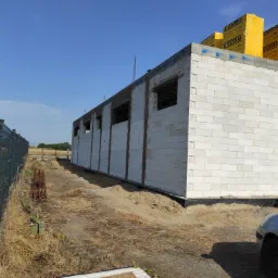 Wznoszenie ścian domu z białych bloczków Ytong, widoczne otwory okienne i palety bloczków na górze, teren budowy ogrodzony siatką.