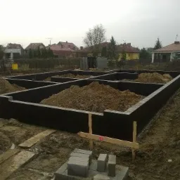 Czarna izolacja fundamentów w kształcie prostokątnych komór wypełnionych ziemią, z widocznymi bloczkami betonowymi w rogu i drewnianymi kołkami wyznaczającymi obrys budynku na tle zabudowy...