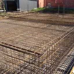 Zbrojenie płyty fundamentowej z widoczną konstrukcją szalunkową i wystającymi prętami pionowymi, przygotowane do zalania betonem.