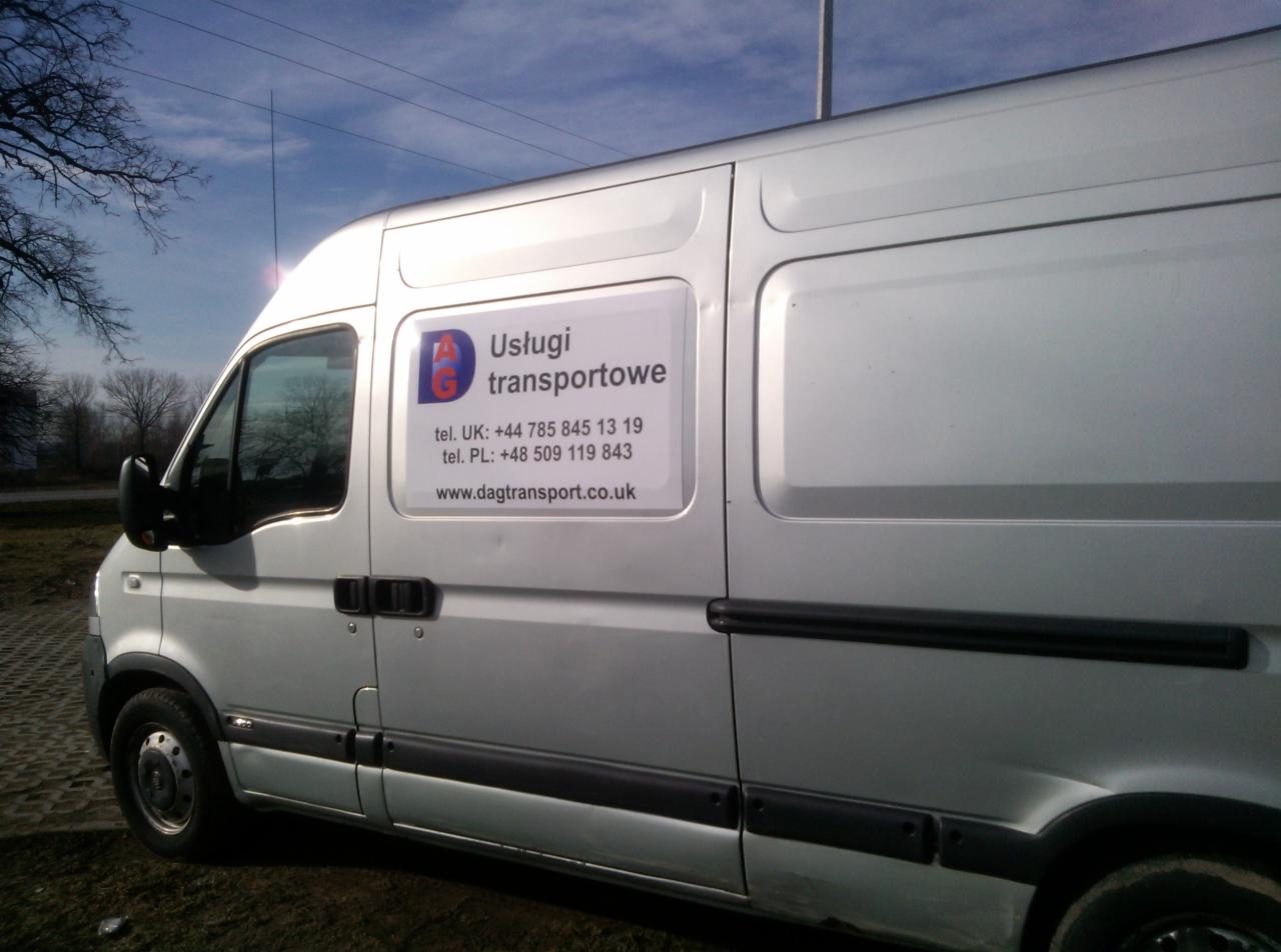 Srebrny bus z reklamą usług transportowych DAG, z numerami telefonów do UK i PL oraz adresem strony internetowej, zaparkowany na trawiastym podłożu.