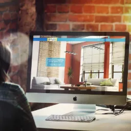 Kobieta przegląda stronę internetową salonu z roletami na iMacu. Widoczny fragment biurka z klawiaturą, tło z cegły.