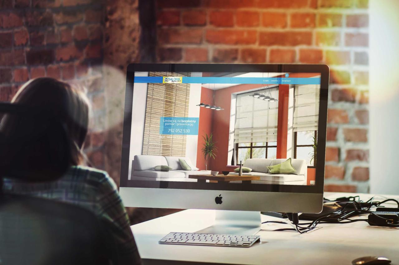 Kobieta przegląda stronę internetową salonu z roletami na iMacu. Widoczny fragment biurka z klawiaturą, tło z cegły.