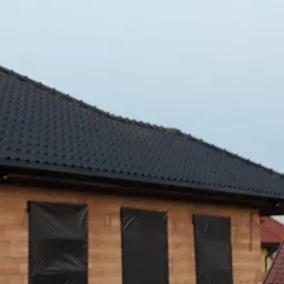 Fragment domu w budowie z czarnym dachem z dachówki ceramicznej i oknami zabezpieczonymi folią, widoczne elementy konstrukcyjne.