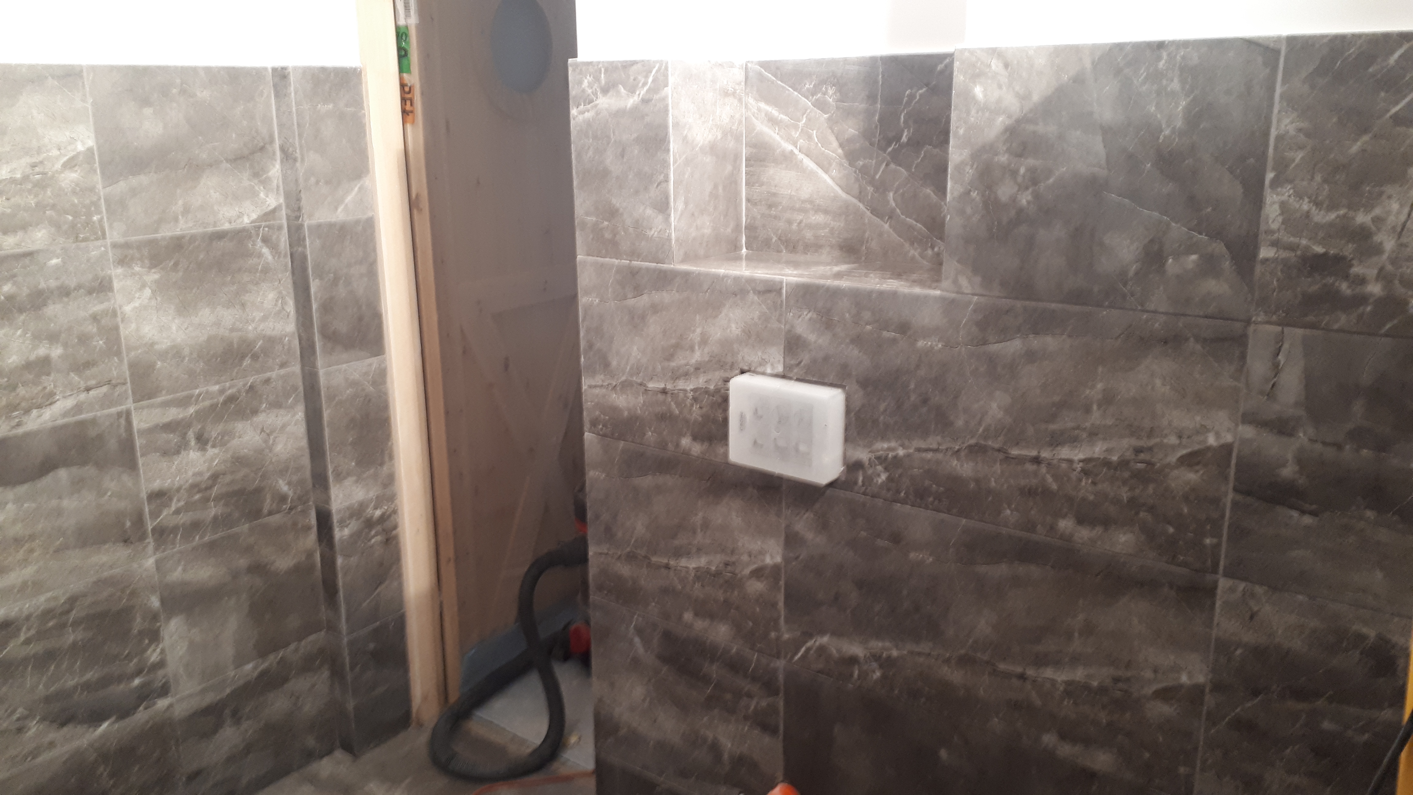 Ściana łazienki wyłożona szarymi płytkami imitującymi marmur, z zamontowanym białym dozownikiem mydła w trakcie wykańczania, widoczne narzędzia i niedokończone obramowanie drzwi.