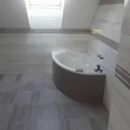 Wykończona łazienka na poddaszu z oknem dachowym, szare płytki podłogowe i ścienne z dekoracyjnym pasem, zabudowana wanna z mozaiką, zabezpieczona folią.