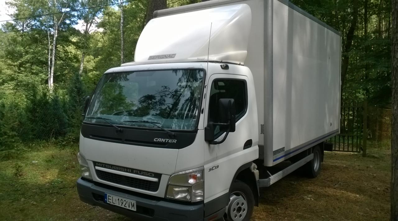 Biały samochód dostawczy Mitsubishi Fuso Canter z nadbudową Serignac, zaparkowany na trawiastym terenie, otoczony drzewami. Widoczna tablica rejestracyjna EL 192VM.