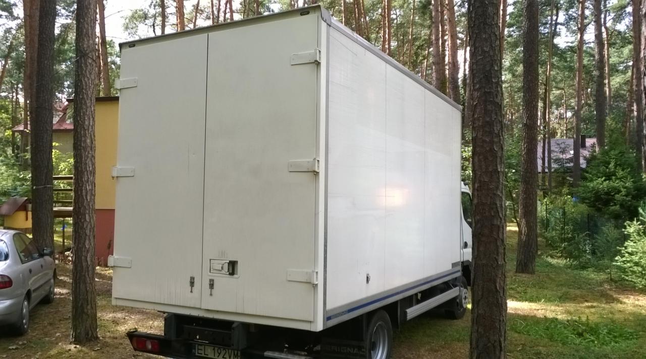 Biały bus dostawczy z zamkniętymi drzwiami, zaparkowany na trawie w otoczeniu drzew. Widok z tyłu pojazdu, tablica rejestracyjna widoczna.