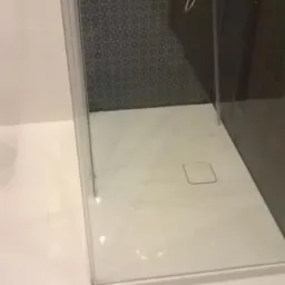 Nowoczesna kabina prysznicowa z białym brodzikiem i czarną mozaiką na ścianie. Widoczny panel prysznicowy ze stali nierdzewnej i szklane drzwi z chromowanymi okuciami.