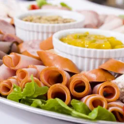 Elegancki półmisek wędlin i serów z dodatkami, w tym musztarda i sos chrzanowy, dekorowany świeżą zieleniną i papryką. Idealny na catering firmowy lub świąteczny.